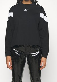 Zwarte sweatshirt met witte schouderaccenten en Puma-logo, met een V-vormige naad, lange mouwen en een cropped fit. Gecombineerd met glanzende zwarte pantalon.