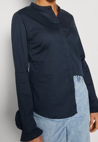 Blusa azul marino de botones con un cuello alto y mangas largas, con puños con volantes y una textura suave, combinada con jeans de lavado claro.