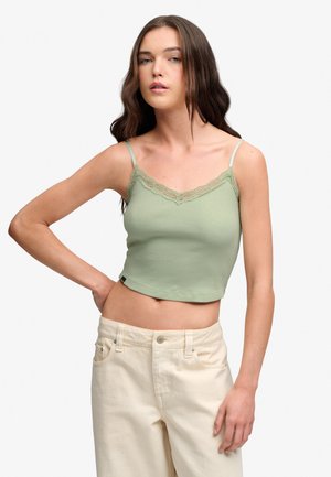 Ung kvinde med langt brunt hår iført en lys grøn crop top med blonder og off-white højtaljede bukser, poserende med den ene hånd på hoften.