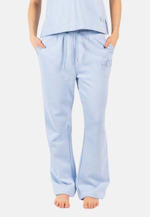 Person som har på seg lyseblå, løstsittende joggebukser med snor og "HÉST"-logo på låret, står barbent med hendene i lommene.