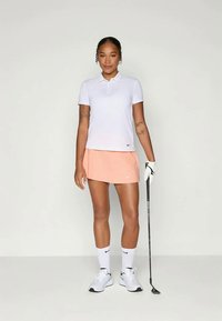 Fehér póló és világos rózsaszín szoknya logóval, fehér zoknikkal és cipőkkel párosítva. Golfütőt tart, egy sima háttér előtt áll.