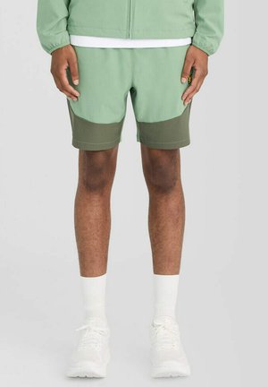 Man draagt tweekleurige groene korte broek, lichtgroene jas, witte sokken en witte sneakers, staand tegen een effen achtergrond.