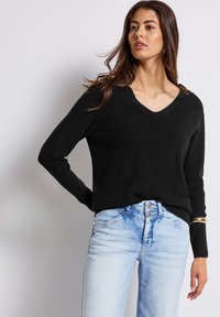 Svart ribbad tröja med V-ringning, tillsammans med ljusblå denimjeans. Tröjan har långa ärmar och en avslappnad passform.