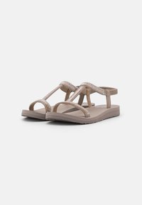 Tan sandals with a geometric strap design featuring small metallic accents, a flat sole, and an open-toe construction. Lightweight material.