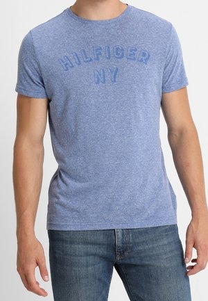 Man in een blauw T-shirt met korte mouwen met de tekst "HILFIGER NY" en een medium-wash blauwe spijkerbroek, staande tegen een effen achtergrond.