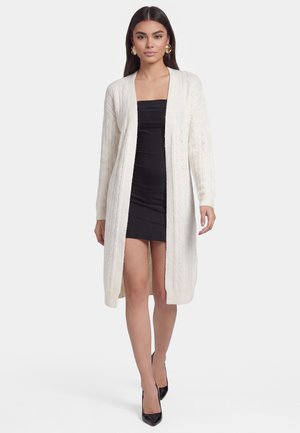 Kvinde iført en hvid lang kabelstrikket cardigan over en kort sort stropløs kjole og sorte spidse høje hæle, står foran en hvid baggrund.