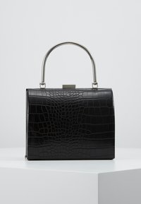Musta suorakaiteen muotoinen käsilaukku krokotiilikuviolla ja hopeanvärisellä kaarevalla metallisankalla, asetettuna valkoiselle pinnalle vaaleanharmaata taustaa vasten.
