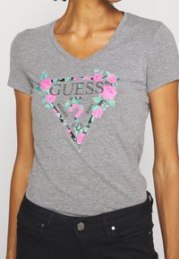 Kvinde iført en grå V-hals T-shirt med et blomstret Guess-logo og sorte bukser, vist fra halsen til hofterne mod en ensfarvet baggrund.