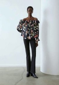 Μπλούζα με γ floral τύπου off-the-shoulder σε σκούρο ύφασμα με φουσκωτές άκρες, συνδυασμένη με εφαρμοστά μαύρα παντελόνια και μποτάκια μέχρι τον αστράγαλο, σε φωτεινό φόντο.