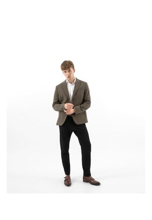 Giovane uomo che sistema un blazer marrone, indossando una camicia bianca, pantaloni neri e scarpe eleganti marroni, in piedi contro uno sfondo bianco.