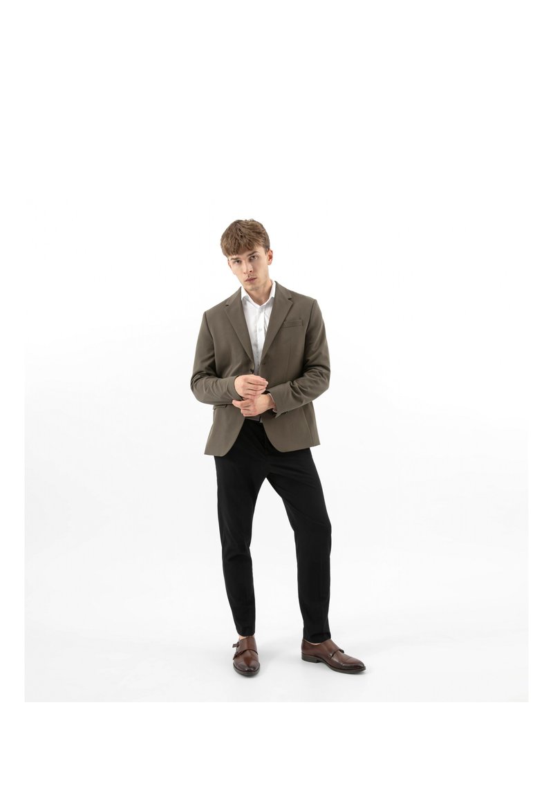 Giovane uomo che sistema un blazer marrone, indossando una camicia bianca, pantaloni neri e scarpe eleganti marroni, in piedi contro uno sfondo bianco.