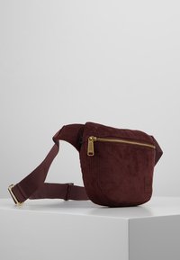 Burgundowa torba na biodra z korduroy z złotym zamkiem błyskawicznym i regulowanym paskiem, wyeksponowana na białej powierzchni na tle prostego szarego tła.