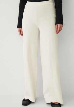 Kvinde iført højtaljede, viddebenede cremefarvede strikkede bukser og sorte sko med rund tå, stående op ad en ensfarvet lys væg.