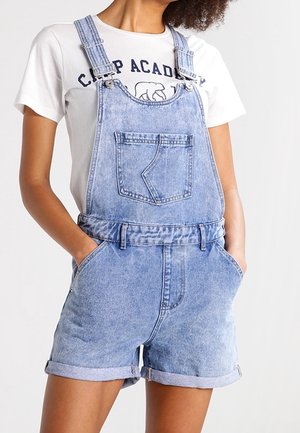Woman wearing light blue denim short overalls with front chest pocket and rolled hem, paired with white t-shirt with graphic text and bear.