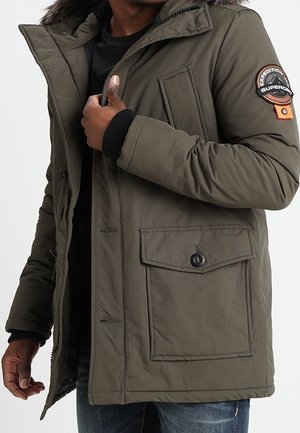 Homme portant une parka vert olive avec capuche bordée de fourrure, grande poche avant et patch circulaire sur la manche supérieure.