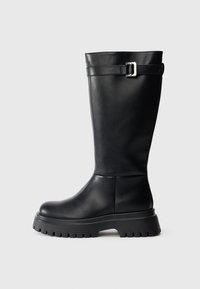 Bota alta preta de couro, com bico arredondado e sola de borracha robusta. Apresenta uma tira com uma fivela retangular perto do topo.