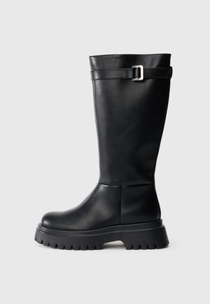 Čierne kožené vysoké čižmy až po kolená s guľatou špičkou a robustnou gumovou podrážkou. Obsahujú popruh s obdĺžnikovou prackou pri vrchu.