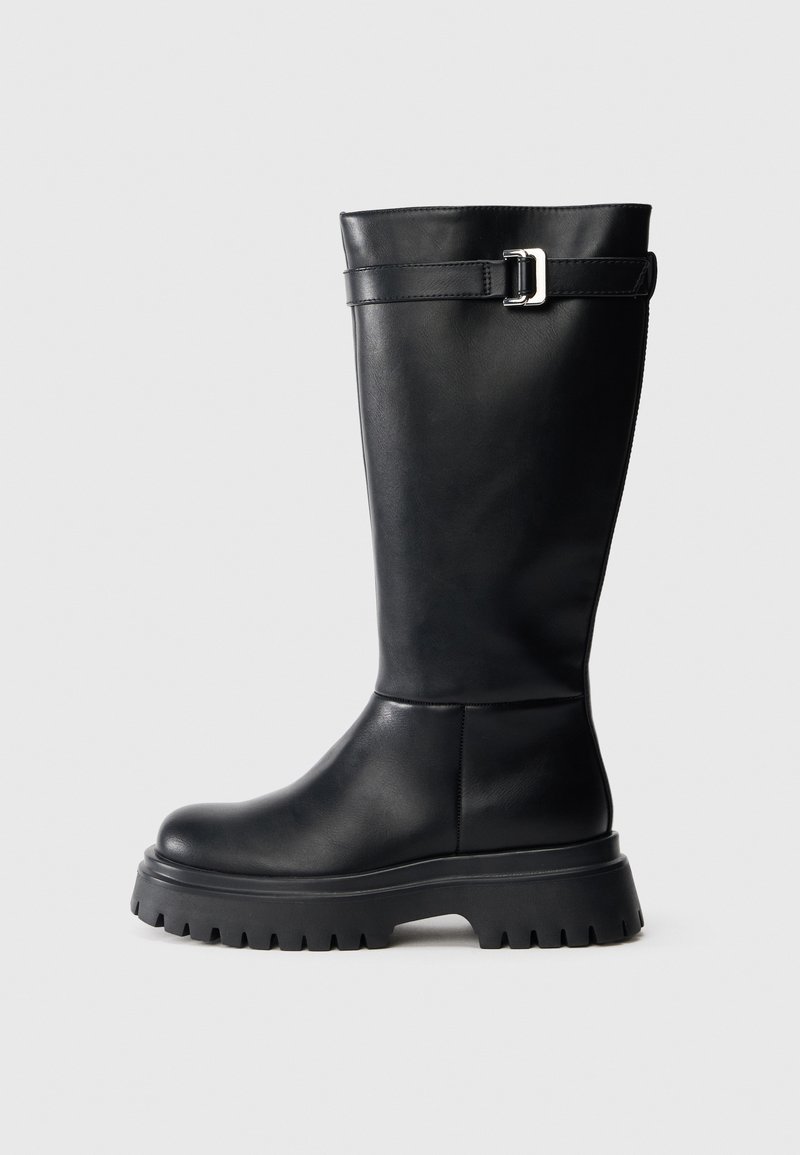 Bota alta preta de couro, com bico arredondado e sola de borracha robusta. Apresenta uma tira com uma fivela retangular perto do topo.