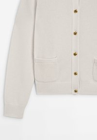 Vaalean beige neuletakki, jossa on ribbadut helma ja hihat. Viisi kultaisia solmupainikkeita ja kaksi etutaskua, joissa on tekstuuripinta.