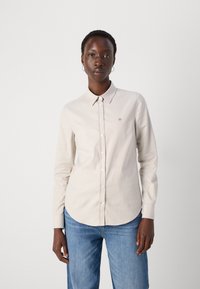Light beige button-up shirt with a collar, made from smooth fabric. Features a small embroidered logo on the chest and long sleeves.