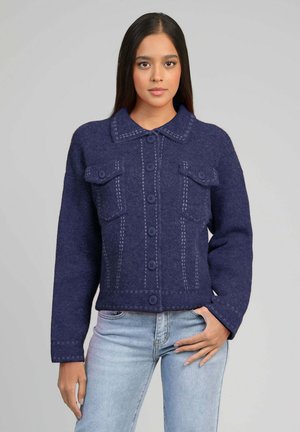 Femme aux longs cheveux foncés portant une veste en laine bleu marine boutonnée avec des poches poitrine et un jean bleu clair, debout devant un fond uni.