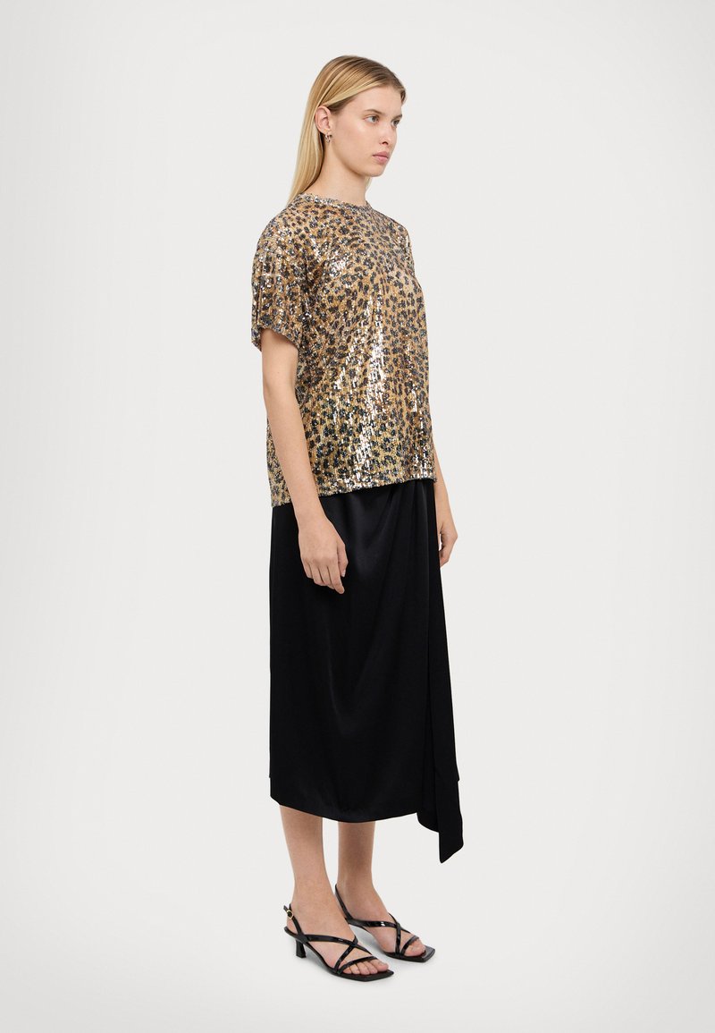 Top in paillettes leopardato oro e nero con maniche corte, abbinato a una gonna nera con un design drappeggiato. Ambientato su uno sfondo neutro.