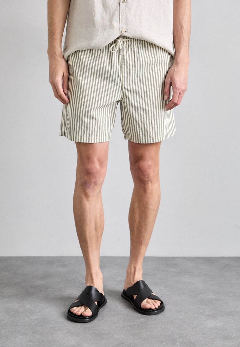 Pantaloni corti leggeri a righe in verde spento e crema, con vita a cordoncino elastico; abbinati a sandali neri con incrocio delle cinghie.