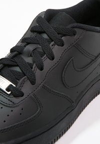 Černé kožené tenisky s texturovaným povrchem, gumovou podrážkou, černými tkaničkami a kovovým štítkem na tkaničkách. Obsahují perforace a vyražené logo.