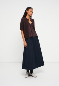 Mulher vestindo um cardigan castanho de mangas curtas sobre um top de alças escuro, saia maxi plissada azul-marinho, meias pretas e mocassins brancos, em pé sobre um chão branco.