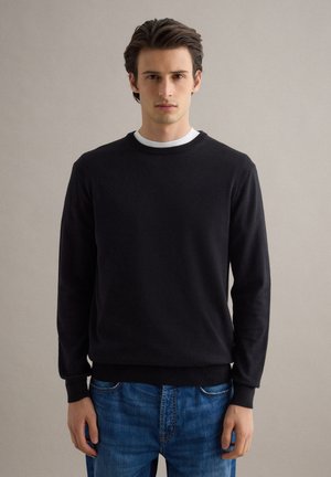 Mann trägt schwarzen Pullover über weißem Hemd und blauen Jeans, steht vor einfarbig hellem grauem Hintergrund und blickt nach vorne.