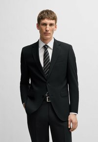 Giacca nera con revers a punta, un bottone, due tasche frontali, indossata sopra una camicia bianca e una cravatta a righe nere, con una fibbia della cintura argentata.