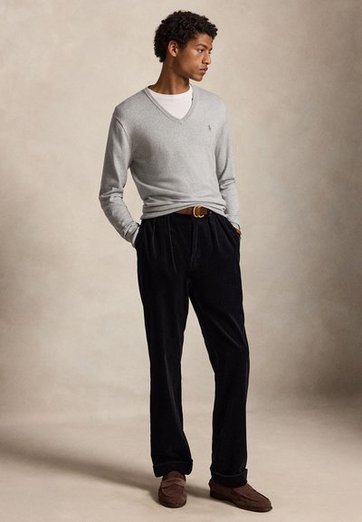 Jonge man staat met handen in de zakken, draagt een lichtgrijze V-hals trui, wit overhemd, donkere broek, bruine riem en bruine loafers.
