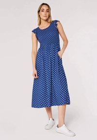 Blue midi dress with white polka dots, short ruffled sleeves, elastic waist, and side pockets. Paired with white sneakers. Smooth fabric texture.