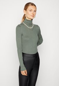 Pullover de gola alta verde com textura à rigas e mangas longas, usado com colares em camadas, combinado com calças pretas de textura suave.