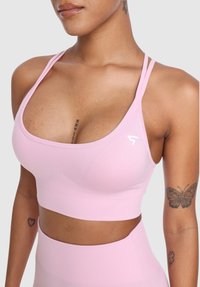 Ženska v svetlo roza športnem modrčku in legginsih, ki prikazuje tetovaže metulja in srca na rokah ter tetovažo rimskih številk na prsih.