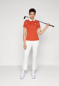 Rotes Poloshirt mit weißen Akzenten, kurze Ärmel, getragen mit weißen Hosen und grauen Sportschuhen. Golfschläger auf der Schulter gehalten.