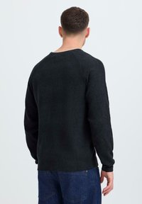 Zwarte ribbeltrui met lange mouwen, ronde halslijn en een gestructureerde afwerking, gedragen met blauwe denim jeans. Achteraanzicht getoond.