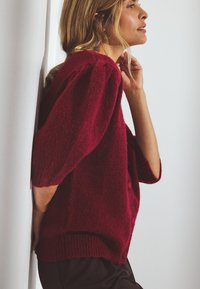 Bordo megztinis su tekstūru, per didelėmis rankovėmis, mygtukais ir raišteliais, dėvimas virš tamsių kelnių priešais sieną.