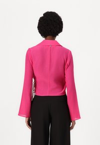 Fuchsia cropped blouse with a collar, long bell sleeves, textured fabric, and a back design featuring a small fastening detail.