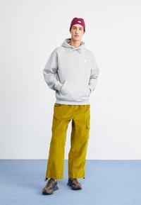 Grauer Kapuzenpullover, gelbe Cordhosen und braune Camo-Sneaker. Der Kapuzenpullover hat eine Fronttasche; die Hose hat Seitentaschen und eine lockere Passform.