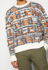 Strickpullover mit strukturiertem Design, der eine weiße Basis und mehrfarbige Muster in Schwarz, Orange und Grau aufweist, mit fransigen Details und gerippten Bündchen.