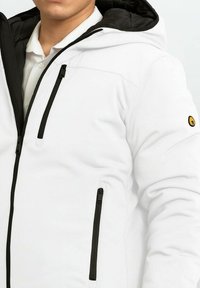 Giacca isolante bianca con dettagli neri, dotata di zip frontale e tasche, cappuccio e una finitura testurizzata per la durabilità.