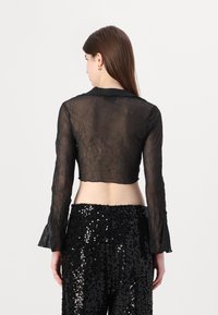 Svart, kort, gjennomsiktig bluse med lange, utsvingte ermer. Stoffet har en krøllete tekstur. Båret over svarte bukser dekket med paljetter.
