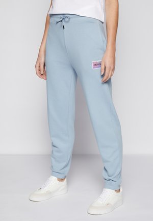 Persoon draagt lichtblauwe joggingbroek met een witte trekkoord en een klein meerkleurig "HUGO" logo op linker dij, witte sneakers.
