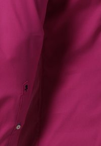 Gros plan d'une manche de chemise magenta avec une petite fleur noire brodée au-dessus d'un bouton blanc.