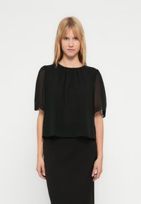 Ξανθιά γυναίκα φοράει μαύρη μπλούζα με διαφανή κοντά μανίκια και κεντήματα με λουλούδια, συνδυασμένη με μαύρη εφαρμοστή φούστα, στέκεται μπροστά σε λευκό φόντο.