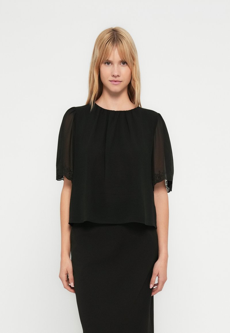 Ξανθιά γυναίκα φοράει μαύρη μπλούζα με διαφανή κοντά μανίκια και κεντήματα με λουλούδια, συνδυασμένη με μαύρη εφαρμοστή φούστα, στέκεται μπροστά σε λευκό φόντο.