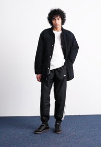 Μαύρο μπουφάν με κούμπωμα, λευκό T-shirt, μαύρη φόρμα με λευκές ρίγες και μαύρα αθλητικά παπούτσια, σε απλό φόντο.