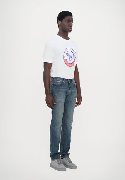 T-shirt blanche avec logo graphique rond, jeans en denim bleu, baskets grises. Design simple, texture douce, silhouette ajustée.