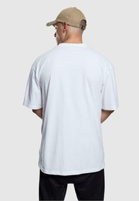 Camiseta de algodón blanca de manga corta, corte relajado, diseño liso, vista desde la parte posterior. Combinada con una gorra de béisbol beige.